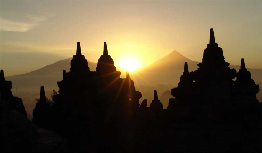Borobodur and Prambanan

Though I love temples I seldom get the chance to  go to temples outside India. So I was very happy to get an invitation from my Japanese daughter to go to Indonesia  to visit the ancient temples of Borobudur and Prambanan.  Thus it happened that I landed in  Jakarta last week and took the next flight to Yogjakarta. This city is also known simply as Jogja or Yogya. This word is actually a corruption of Ayodhya, the famed city of Rama in India. Names from the Ramayana abound in this island. Sometimes even without their knowing it many of the names used by the Indonesians are Sanskrit in origin but of course most of them are so distorted that it takes a while to find out the actual source.  The Special Region where Yogyakarta is situated is the smallest province of Indonesia (excluding Jakarta). It is located on the island of Java. The word Java has such a special flavour that I was really glad that I had come here instead of one of the big cities. On a clear day one can see the famed volcano Mt. Merapi, which is still alive and the history of which is closely connected with the history of Borobudur and Prambanan.  
 This 300 hundred old city is the cultural centre not only of Java, but of the whole of Indonesia. This is in part due to the variety of religious influences found here like, Buddhist, Hindu, and other indigenous beliefs, which resulted in the construction of the impressive temples of Borobudur, Prambanan and the Dieng temple complex. 
Indonesia's oldest palace 'The Kraton,' is situated here. This is still the home of Jogya's royalty. It is the only province in Indonesia that is still formally governed by a Sultanate, the Sultanate of Ngayogyakarta Hadiningrat. Even now the current Sultan of Yogyakarta retains remarkable political prestige. Yogyakarta offers an abundance of Javanese art, painting, silverwork, batik handcraft, traditional Javanese dances, as well as contemporary art. 
From Yogyakarta airport we took a taxi to Borobudur where we stayed at the Manohara hotel which is supposed to be the only one with a magnificent view of the temple which can be seen right in front of it. The hotel is set amidst a typical tropical garden filled with lush greenery and the usual tropical flowers like ixora, frangipani, bouganvilleas in variegated shades, sweet smelling jasmine and the famous paritjata flower which is mentioned in our puranas. There was a unique blend of cultures even in the décor. We were greeted by two ancient Ganesha statues at the entrance but there was a pool in front of the hotel in which you found a statue of Lakshmi which had a striking resemblance to the Chinese goddess of prosperity called Kuan Yin and scarcely any resemblance to the Hindu goddess.  At night four huge water lilies bloomed, shedding their intoxicating fragrance into the air. I had read about these night blooming water lilies called 'kumudini' in Sanskrit literature but it was the first time that I had ever seen them. They were pure white and as big as lotuses. They reached their maximum size at midnight and closed up again by morning.
Watching the sunrise from the top of the temple was supposed to be a great experience but again we were told that since it was the rainy season it was very unlikely that we would be able to see it. I breathed a sigh of relief that I wouldn't have to jump out of bed at 3 in the morning and dash up the hill. I had been traveling continuously from 4 am the previous day. It was a grueling journey from Rishikesh to Delhi and from Delhi to KL and then to Jakarta and to Yogyakarta and finally to Borobudur!!
However we set off at 10 in the morning along with a host of other sight seers. They were certainly not pilgrims and all of them were armed with hats and caps of various sizes and designs and of course the umbrella man was having a great trade selling multi colored umbrellas. So armed with one of these I set off in the blazing hot sun, not waiting for the rest of the party who were frantically searching for a good guide. Since I was a very slow walker and even a slower climber I fully expected the young members of my party to catch up with me in no time. But I climbed and climbed for more than half an hour without seeing any of them. Each time I turned round to have a look, all I could see was a sea of brightly colored umbrellas bobbing up and down. Eventually I gave up the attempt and decided to carry on to the top on my own. I did two of the pradikshanas or circumambulations of the 5th and 6th platforms and finally reached the top to get to which one had to climb some steps which were obviously made for giants. I was totally drenched in perspiration and would have been glad to get at least a tiny bit of shade but there was none to be had since all the pagodas hardly cast any shade as it was 12 noon. As I stood in the blazing hot sun, not knowing whether to go back or wait for the party, a number of college students clustered round me and started to talk to me very eagerly. They were so sweet that despite the heat I stood and answered all their questions. They had some sticky Indonesian sweets which they pressed on me. I was forced to take some even though the thought of eating sticky sweets when my whole body was sticky and hot was not a very happy thought. Before we parted they presented me with a pearl rosary and of course I gave them our ashram cards with the photo of baby Krishna by which they were quite charmed. I realized afterwards that this is typical Indonesian behavior. They are such a charming people, so friendly, so polite and so interested in people from India.  I started my journey back to the ground soon after since I realized that unless I got to a shade soon I would probably collapse from the heat.
Now to describe the temple….  If seen from the air and also from the small reproduction of it in the museum I realized that it was made in the form of a Hindu Meru which is a vertical representation of the Sri Yantra. Apparently this was the shape of a Buddhist mandala. The height of the whole edifice before renovation was 42 meters. Now it is only 34.5 meters  since the lowest level has been used as a supporting base.
 It has ten terraces. The first six terraces are square and the two upper terraces are circular. Right on top is the terrace on which the statue of the Buddha had been seated facing westward. The statue has been removed and is now to be found in the museum. Each terrace symbolizes one of the stages of human life.  The aspirant who wants to reach the stage of the Buddha has to go through each of these stages. The base is known as Kamadhatu. There is a very similar square in the Sree Yantra which is supposed to be that state of life in which the human being is still bound by desires 'Kama' and passions. From here we climb up to the second, third and fourth terraces and these are known as Rupadhatu. This is the stage when the human being is still bound to 'rupa' or form that is to the world of shapes and forms - the world of the senses as we experience it. On these four terraces the effigies of the Buddha are placed in open space. The next three terraces are known as Arupadhatu and here the effigies of the Buddha are confined inside domes with holes in them. Only those who have freed themselves from lust and passion for forms, can reach this stage. The final part is known as Arupa and is the state of nirvana, or liberation which is the state of the Buddha 
Borobudur is said to have been built by King Samaratungga, one of the kings of the old Mataram Kingdom, the descendants of the Sailendra dynasty. Based on Kayumwungan inscription, an Indonesian named Hudaya Kandahjaya had a revelation in which he was told that Borobudur had once been a place for prayer that was supposed to be completed  on 26 May 824, almost one hundred years after the construction had started. The name of Borobudur, as some people say, means a mountain having terraces (budhara), while other says that Borobudur means monastery on a high place. 
It is a  Buddhist temple and has 1460 relief panels and 504 effigies of Buddha in its complex. These panels are all on the walls of the terraces. In order to see them one has to climb the stairs of each of the terraces and then do a pradikshina  or circumambulation in a clockwise direction round it. This is the correct way to discover the beauties of the temple and also derive maximum spiritual benefit.
Each terrace has its own panels showing how skillful the sculptors were. Some of these panels tell the legendary story of the Ramayana. Besides these, there are panels depicting the condition of the society at that time.  Some show farmers working in the fields and some show sailors in boats, thus giving us a glimpse of the advanced state of the navigation techniques of that age.
Many of the panels reflect the Buddha's teachings. If you walk through each narrow passage in Borobudur with a competent guide you will come to know much of the philosophy of Buddhism. Atisha, a Buddhist monk from India in the tenth century once visited this temple that was built 3 centuries before Angkor Wat in Cambodia and 4 centuries before the Grand Cathedrals in Europe. After gathering a lot of information from these reliefs he returned to India and started another sect known as Vikramashila Buddhism. Later he became the leader of the Vikramashila monastery and became a teacher in Tibet. Six scripts from Serlingpa were then summarized as the core of the teaching called "The Lamp for the Path to Enlightenment"  known as Bodhipathapradipa.
The scupture of the panels leaves you spellbound. Great attention is paid to the most intricate details. Some portions show what happens to those who do evil deeds and what happens to those who do good deeds. Buddha's dictum was that karma or action alone decides our life both now and in there hereafter. 'As we sow, so we shall reap.'
Some of the panels depict the 'Jataka Tales' in which moral questions are answered  through beautiful tales of birds and animals.
The mystery of why the temple was constructed in the first place and how it was buried for centuries is still not clear. Some say that initially it was surrounded by swamps and was buried because of the volcanic eruption of Mt. Merapi. 
Sir Stamford Rafles was said to be the one who first discovered the existence of this site and encouraged a German to excavate the huge mound and unearth the wonders of this temple. Since then the Indonesian government as well as the UNESCO has done a lot of restoration on the temple which is still being carried on. However when you go there in the early morning, you can have a glimpse of what it might have been in ancient times. The edifice looms through the pearly mist of a monsoon day and one can imagine the wonder and the mysery of that amazing place and imagne the ancient Buddhist pilgrims plodding their way up with their rosaries swinging from their hands muttering the great mantra of the Buddha - 'Aum Mani Padme Hum'. 
 

PRAMBANANA TEMPLE
We left Borobodur reluctantly and returned to Yogjyakarta.  Our next port of call was Prambanan which is a ten-century Hindu temple. This temple is dedicated to Shiva, and is locally known as Candi Loro Jonggrang, which means 'temple of the slender virgin.' The beautifully sculptured spire, of the main temple which soars up to a height of fifty meters  does indeed resemble a 'slender virgin.' Like the Buddhist stupa in Borobudur , Prambanan was probably abandoned when the Buddhist and Hindu inhabitants of Java moved to East Java. The temple is situated seventeen kilometers east of Yogyakarta, and is believed to have been built by King Balitung Maha Sambu in the middle of the ninth century. Another story goes that it was built around 850 CE either by Rakai Pakatan of the second Mataram dynasty, or Balitun Maha Shambu, during the Sanjaya Dynasty. Not long after its construction, the temple was abandoned and began to deteriorate. This was no doubt due to some fault in the location of the site according to Vaastu - the Indian science of architecture.Reconstruction of the compound began in 1918. The main building was completed around 1953. Much of the original stonework had been stolen and used at other sites. A temple will only be rebuilt if at least 75% of the original stones are available, and therefore only the foundation walls of most of the smaller shrines are now visible and there are no plans for their reconstruction.
The temple was again severely damaged during the earthquake in Java in 2006. Early photos suggest that although the complex appears to be structurally intact, damage is significant. Large pieces of debris, including carvings, were scattered all over the ground. The temple was closed to the public for  quite a while. However, some weeks later in the same year the site re-opened for visitors.
The temple complex is set like a jewel amongst luxuriant green fields and some villages. It has eight shrines, of which the three main ones are dedicated to the trimurtis, Shiva, Vishnu and Brahma. The main temple of Shiva rises to a height of 130 feet and houses the magnificent statue of Shiva's consort, Durga. 
The whole complex consists of three zones. The outer zone is a large space marked by a rectangular wall which has been destroyed. It is possible that it was once a sacred park, or an ashram for the priests. In fact the very word Prambanan is actually a corrupt version of the Sanskrit word 'Brahmana' or priest belonging to the Brahmin caste. No doubt these were the ones who came from India and made this temple to their favouite deities.
The middle zone consists of four rows of 224 individual small shrines. These concentric rows of temples have identical designs. Each row is slightly elevated towards the center. These shrines are called "Candi Perwara" or complementary temples. The Perwara are arranged in four rows around the central temples. Perhaps the Brahmin priests made these differentiations according to the four castes and each caste had a special enclosure. The enclosure next to the central compound was accessible to the Brahmin priests only, the next was reserved for the nobles or the Kshatriyas, and the third and fourth for the Vaisyas and Shudras. The very name of the temple suggests that it was a Brahmin controlled hierachy. 
The central compound was no doubt the holiest of the three zones. It has a square elevated platform surrounded by square stone walls with stone gates on each of the four cardinal points. This is surrounded by eight main shrines called candi. The three main shrines as has been mentioned are dedicated to the Trimurtis or Brahma, the Creator, Vishnu, the preserver and Shiva, the destroyer. The three shrines in front of these main temples are dedicated to vehicles of the three gods. The shrine in front of the Brahma temple is to Hamsa or the swan which is Brahma's vehicle, the one in front of Shiva is to Nandi or the bull and before Vishnu we have the Garuda or the eagle vehicle of Vishnu.
 The Shiva shrine at the center contains five chambers. The four small chambers face the four cardinal directions and the main sanctum is reserved for the deity. It used to have a  three meter high statue of Shiva as Mahadeva. The statue of Shiva stands on the yoni or pedestal that bears the carving of  serpents on the north side. The other three smaller chambers contain statues of Shiva's consort Durga,, the sage  Agastya, and Ganesha, the son of Shiva.. Agastya occupies the south chamber, and Ganesha the west, while the north chamber contains the statue of Durga as Mahisasuramardini depicting her slaying the buffalo demon. The shrine of Durga is also called the temple of Lara Jonggrang (Javanese: slender virgin), after a Javanese legend of princess Lara Jonggrang.
This is a very popular legend which connects the site with the Ratu Boko Palace. The legend goes of how Prince Bandung Bondowoso fell in love with Princess Lara Jonggrang, the daughter of King Boko. But the princess rejected his proposal because Bandung Bondowoso had killed King Boko and taken over her kingdom. Bandung Bondowoso forced her to agree ro marry him, but she set him one condition. The Prince would have to build her a thousand  temples in one night.
The Prince went into meditation and conjured up a multitude of spirits  from the earth. Helped by these supernatural beings, he succeeded in building 999 temples. When the prince was about to complete the thousandth, the princess woke up her palace maids and ordered the women of the village to begin pounding rice and to make a fire to the east of the temple, in order to make the prince and the spirits believe that the sun was about to rise. As the cocks began to crow, the supernatural helpers fled back into the ground. The prince was furious about this trick and cursed Lara Jonggrang to turn into stone. And thus alas the poor unfortunate princess became the last and the most beautiful of the thousand temples. According to tradition, the Shiva temple is the unfinished thousandth temple created by the demons and the Princess became the image of Durga in the north cell of the temple and thus it came to be known as Lara Jonggrang or Slender Virgin.
The bas-reliefs along the balustrades of the gallery around the Shiva and Brahma temples depict the Ramayana legend. They show how Sita who is known as Sinta in Javanese was abducted by Ravana, the demon king. The monkey king Hanuman brings his army to help Rama and rescue Sita. This story is also shown by the Ramayana Ballet which is regularly performed at full moon at the Trimurti open air theatre in west side of the illuminated Prambanan complex. On the balsutrades in Vishnu temple there is series of bas-reliefs depicting the story of baby Krishna which they call the Krishnayana., which is a word which is never found in India.
That night we went for the Ramayana ballet which was performed on an open air stage with a beautiful background . The costumes of the women reminded me of the costumes of dancers or Odissi. The villains make up was reminiscent of the Kerala Kathakali. All the names had strange twists in them and even the story  had some things which are not to be found in the original Ramayana. Hanuman looked very fierce but Rama and Shinta were very sweet. It was a beautiful experience and a wonderful finale to my trip to Indonesia.
Borburdur
